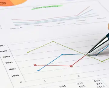 A close-up of a hand holding a pen pointing to a line graph on a document, surrounded by charts and data.